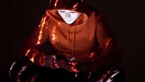 a faceless hacker in an orange hoodie is illuminated by red code reflections while using a smartphone in the dark, highlighting covert digital activity and mobile-based cyber interference
