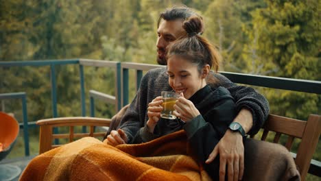 The-girl-is-sitting-on-the-couch-next-to-her-brunette-boyfriend-and-drinking-tea,-wrapped-in-a-blanket.-Relax-in-a-country-house-with-a-view-of-the-mountains-and-the-forest