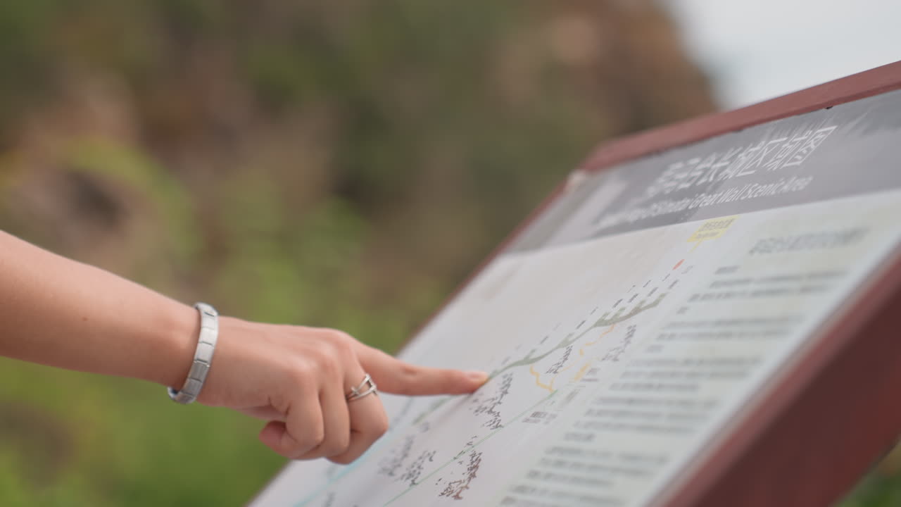 Close up hand points at map with fingertip, silver bracelet visible, detailed map text and route lines in focus, outdoor greenery blurred in background, careful map reading and route confirmation