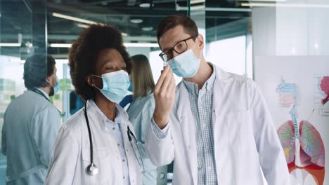 médicos afroamericanos y caucásicos, hombres y mujeres, con máscaras médicas, de pie en el consultorio del hospital, discutiendo sobre la vacuna contra el coronavirus