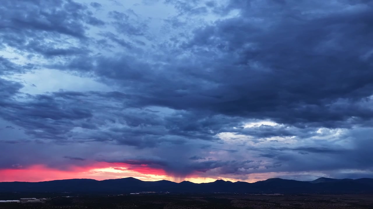 Timelapse 4K con dron sobre el condado de Los Álamos capturando un cielo sombrío y tormentoso sobre las montañas Jemez. Un destello dramático e intenso de luz roja se desplaza por el horizonte bajo densas nubes azules.
