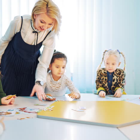 children learn at round table with smiling teacher in class. little kids taking cards to themselves from table during english lesson indoors.