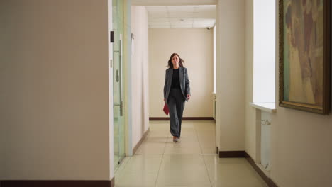 confident businesswoman in formal suit walking down bright office hallway holding red folder, heading to meeting or presentation with purposeful stride in professional corporate environment