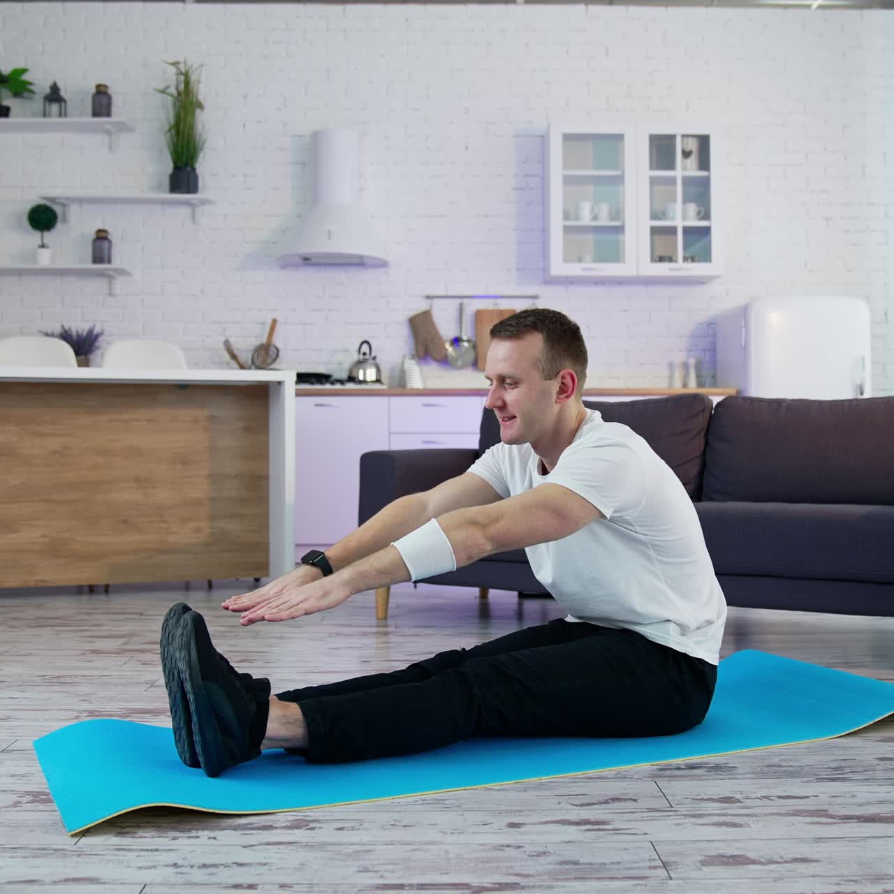 Young man exercising at home. Man sitting on a mat and trying to touch his feet on kitchen background. Difficulties of physical training during lockdown period