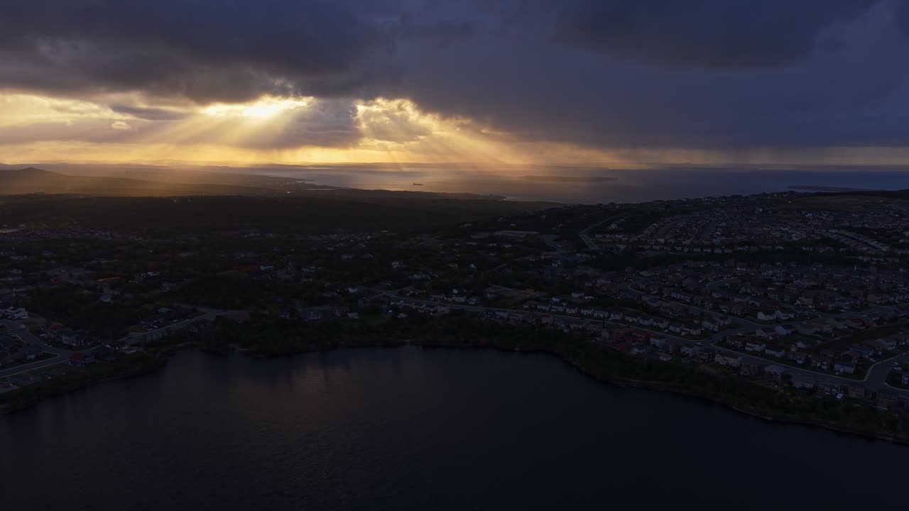 Aerial footage of Conception Bay at dusk where dramatic godrays pierce heavy clouds to illuminate dark waters, creating a moody yet serene atmosphere above Newfoundland