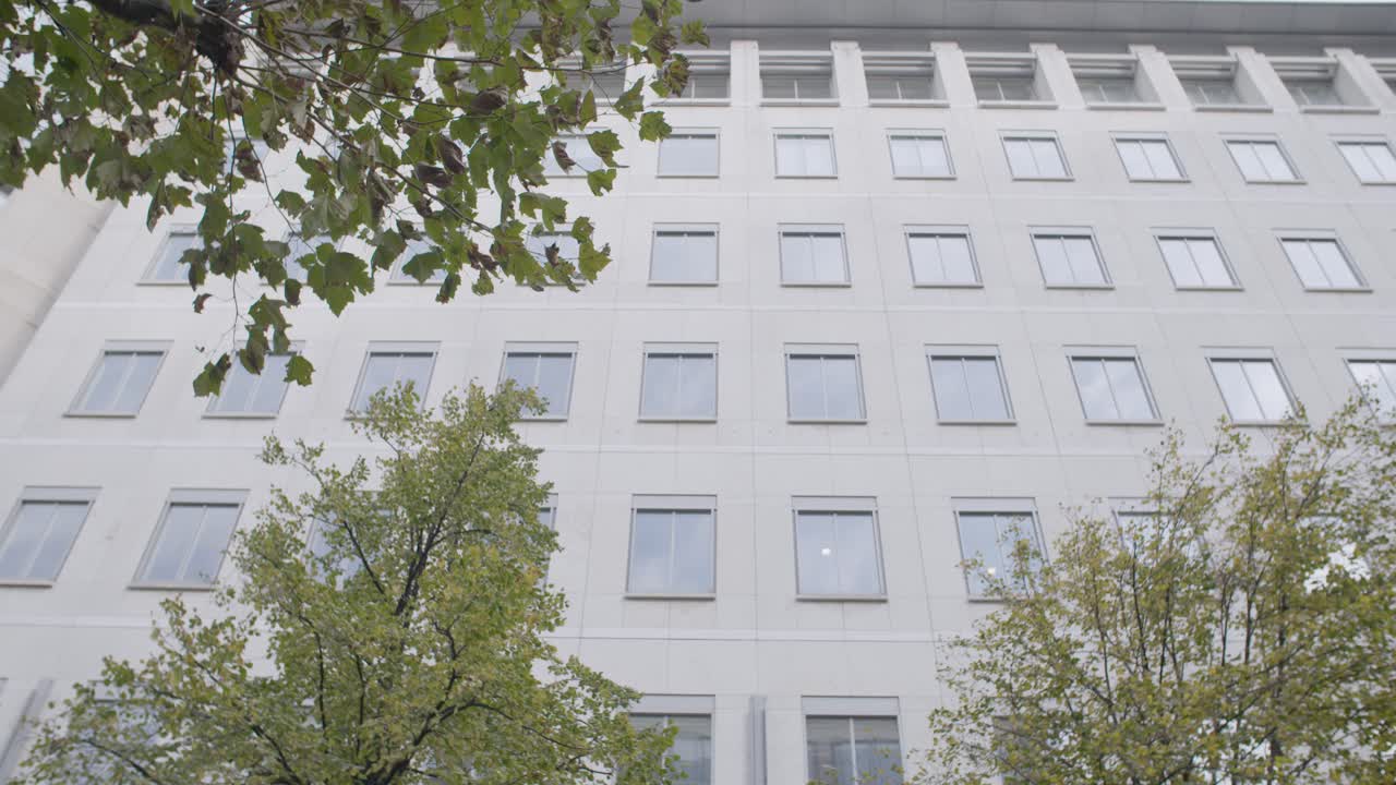 People walk past the entrance of a modern office building surrounded by trees, tilt down shot