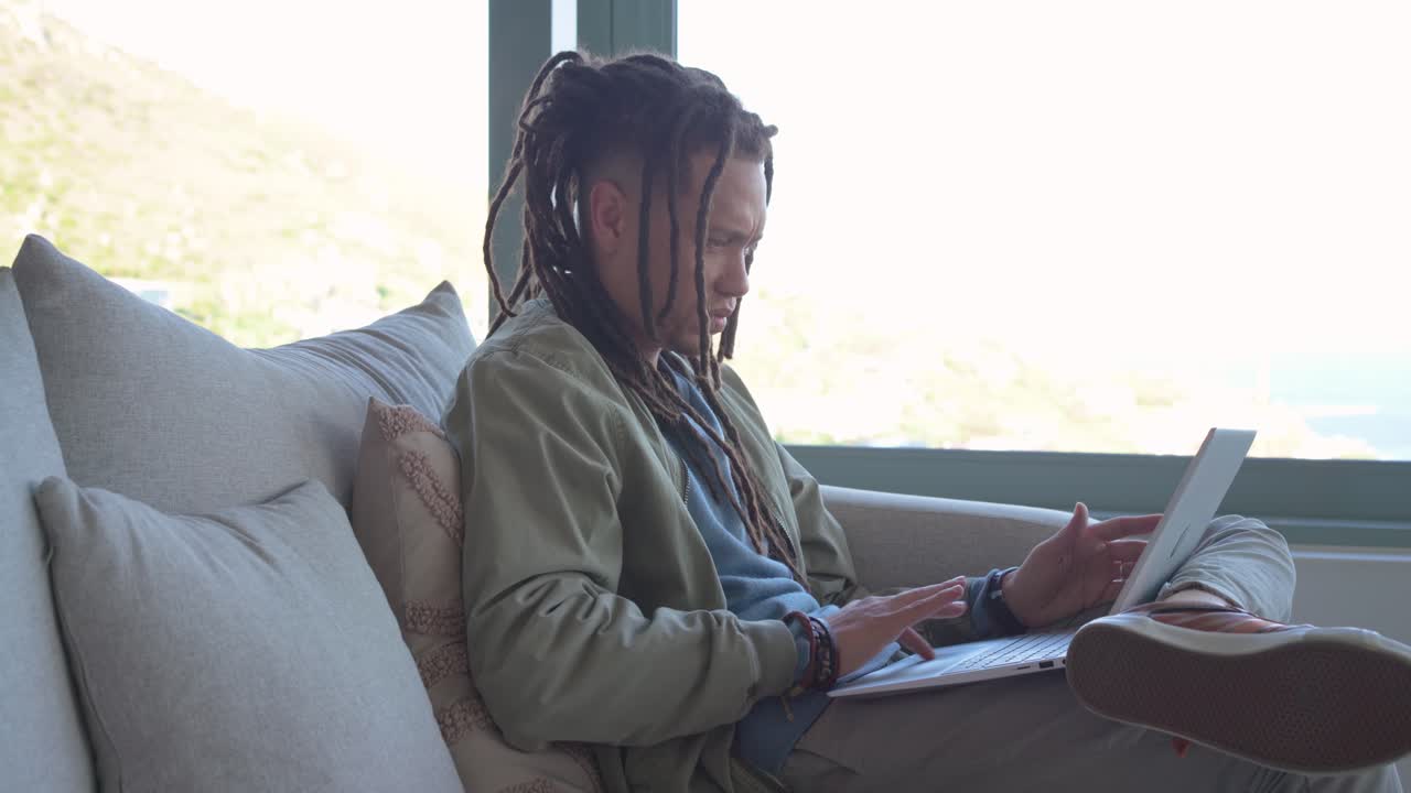 Man typing on laptop on couch by window, adjusting posture and tilting screen for remote work