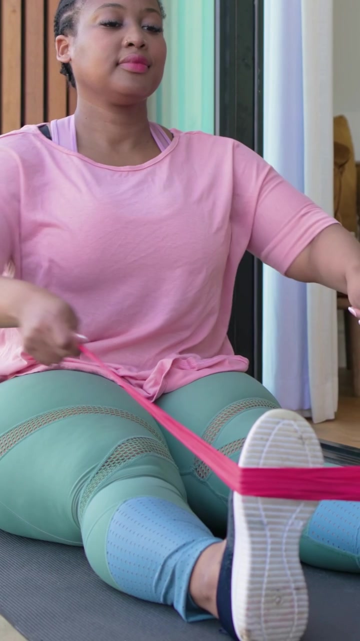 Vertical video: Placing pink band around right foot, woman in leggings rowing for strength at patio