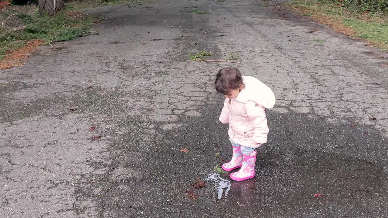 A little girl jumping and dancing in a puddle wearing pink rubber boots ...