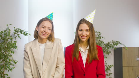 two happy female colleagues with party hat hugging and looking at camera ath the office party