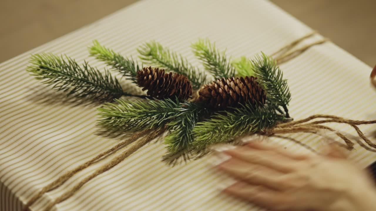 A Woman Wrapping A Christmas Gift Adorned With Evergreen Foliage And A Pine Cone. High Angle Shot