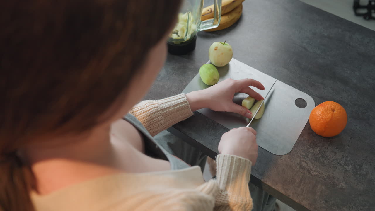 Donna incinta che affetta una pera sul bancone, futura mamma che cucina in una cucina accogliente, primo piano delle mani che tagliano spicchi, manica di maglione di lana, arancia nelle vicinanze, luce naturale del mattino, preparazione di uno spuntino sano per la gravidanza.