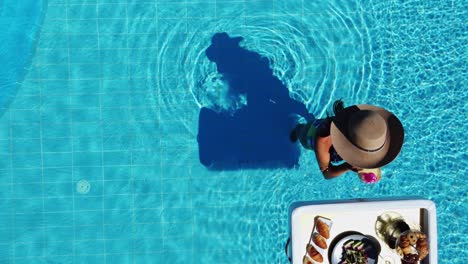 Hermosa-Mujer-Con-Sombrero-Disfrutando-De-Una-Merienda-Y-Una-Bebida-En-Agua-Fría-En-La-Piscina