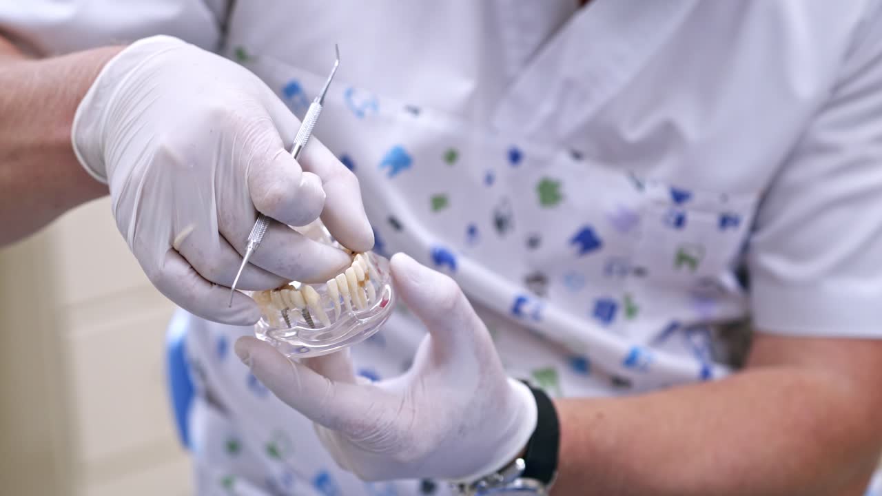Doctor shows on a plastic jaw sample or model different methods of teeth treatment. Modern dental clinic background. Health concept. White medical gloves on male doctor's hands.