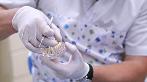 doctor shows on a plastic jaw sample or model different methods of teeth treatment. modern dental clinic background. health concept. white medical gloves on male doctor's hands.