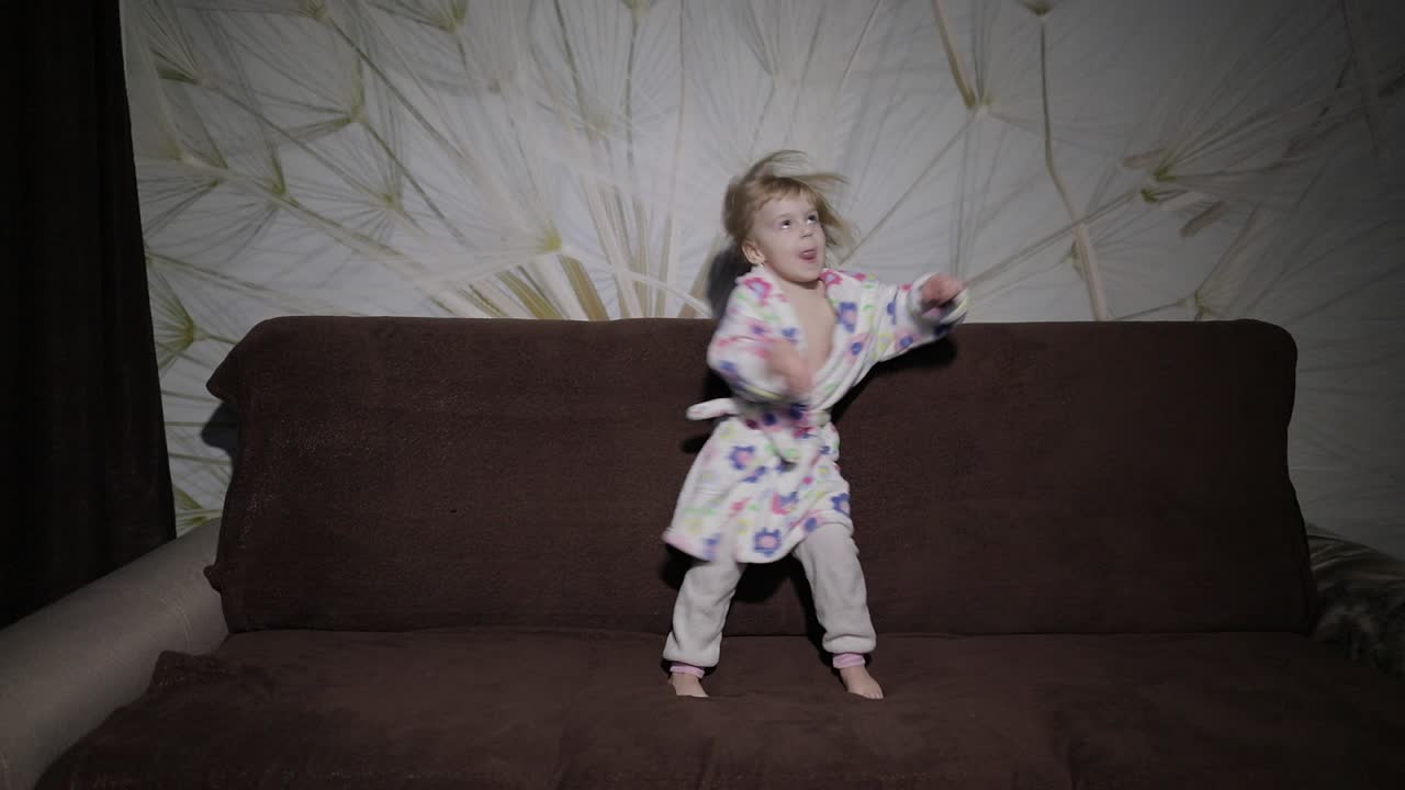 Little cute girl with blond hair jumps on sofa. Bathrobe clothes. Happy