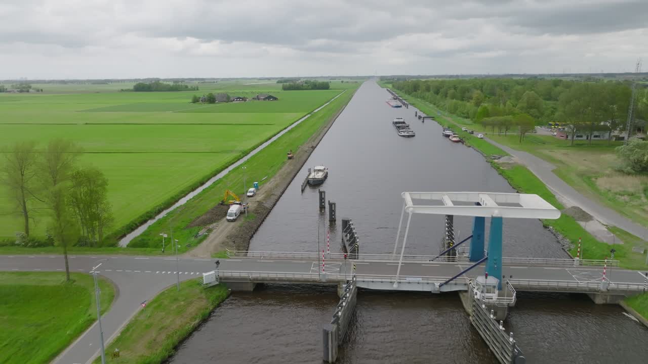 Long perspective down a canal with a large tanker barge passing moorings toward a bascule bridge in open fields