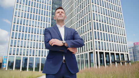 caucasian business man straightens suit and crosses arms by skyscraper