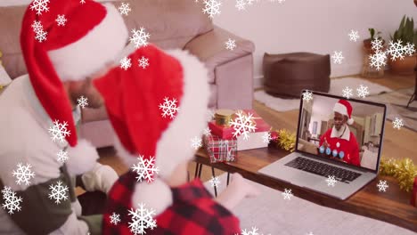 Animation-of-snow-falling-over-smiling-father-and-son-in-santa-hats-on-laptop-video-call-with-family
