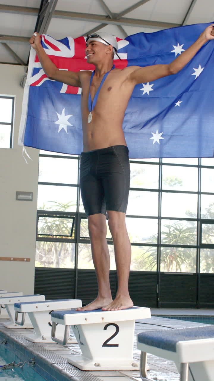 Vertical video: Standing on podium, swimmer holding Australian flag and wearing medal in indoor pool