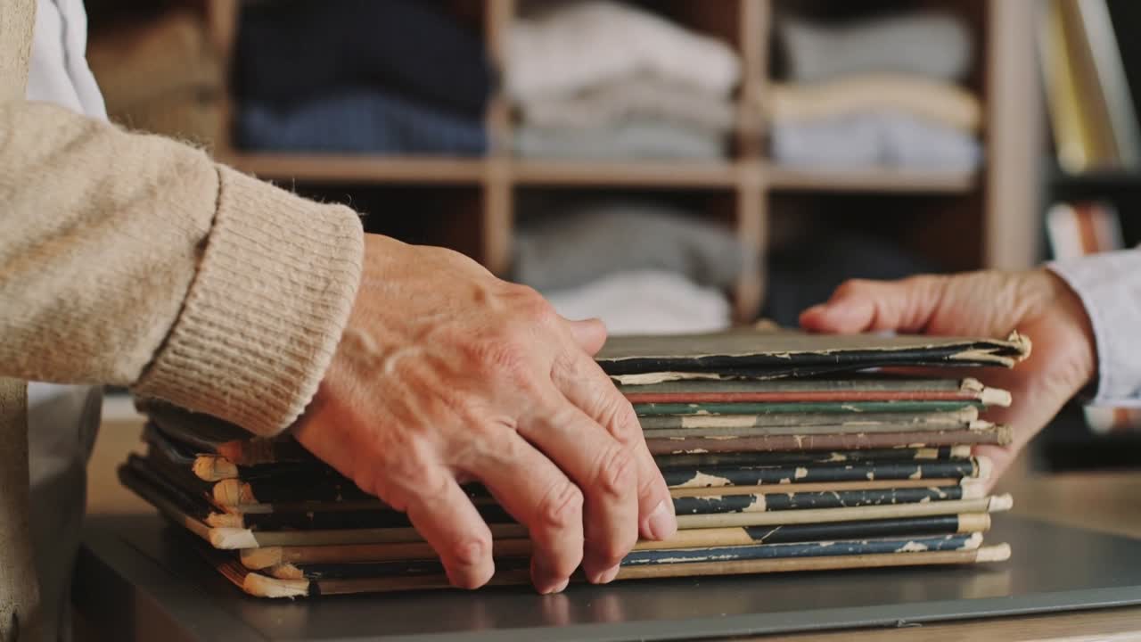 Slidte vinylcovers rækkes over en butiksdisk af en højre hånd, mens en venstre hånd i beige strik arrangerer dem til salg.
