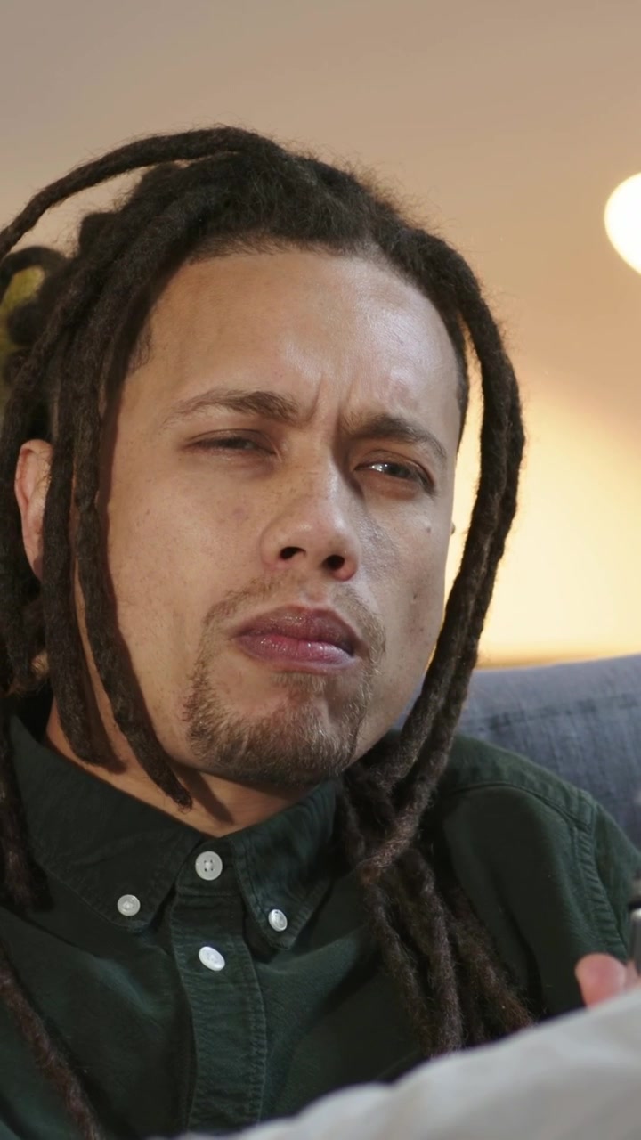 Vertical video: Sipping drink, man with dreads in green shirt pressing remote, adjusting TV at home