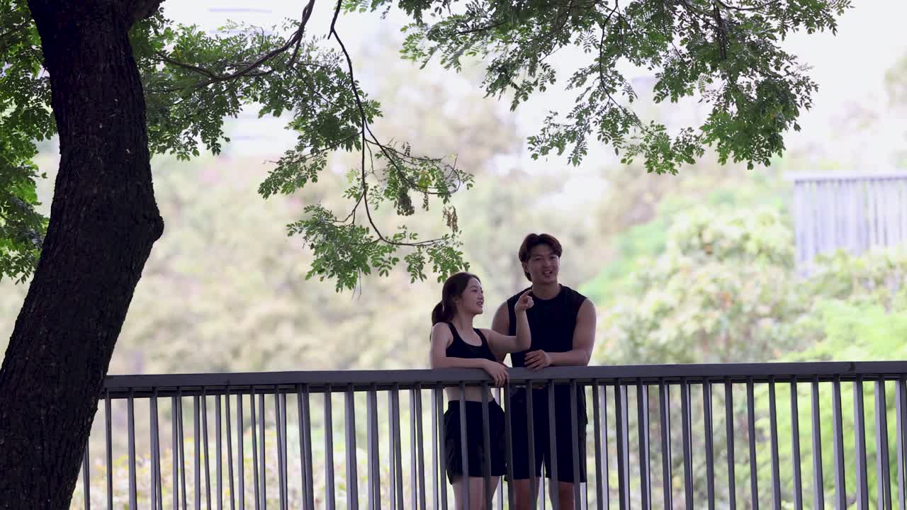 คู่รักชาวไทยวัยหนุ่มสาวในชุดออกกำลังกายยืนคุยกันอยู่บนสะพานในสวนสาธารณะ
