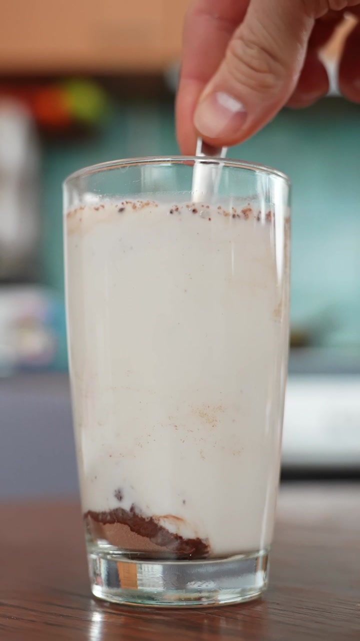 Vertical close-up of a hand swirling homemade chocolate milk in a tall glass on a wooden table, creamy froth with cocoa specks, soft kitchen background, copy space.