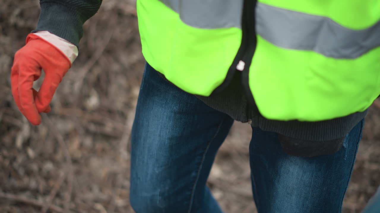 Volunteer wearing reflective vest and gloves picking up discarded plastic bottle from ground covered with dry leaves and twigs during environmental cleanup to protect nature and reduce pollution