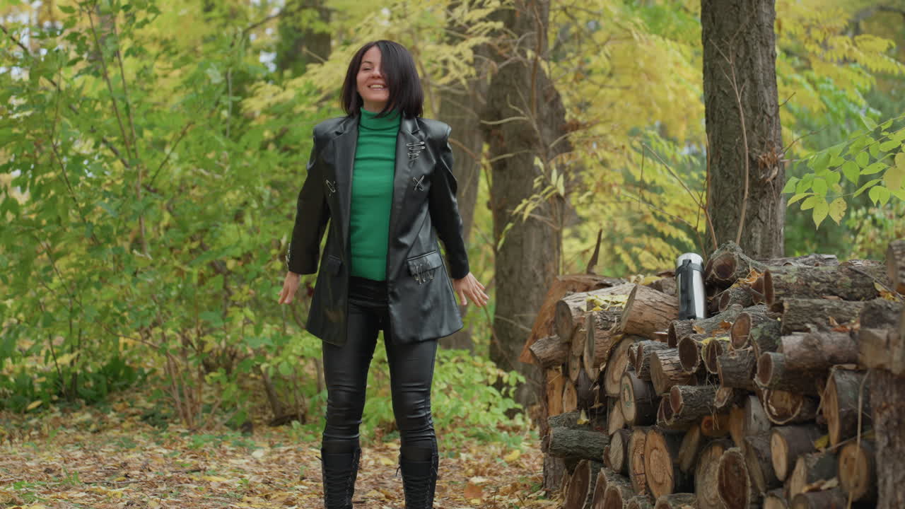 Woman in leather jacket standing among autumn trees, green sweater, black boots, leather bag, stacked logs with thermos, leaves covering ground, confident pause surveying woodland path, cool overcast