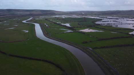 Aerial-Drone-Landscape-of-Water-Channel-Flowing-through-green-agricultural-field-below-small-hills,-wet-parcels,-morning-skyline