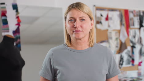 tall beautiful dressmaker smiling warmly while looking directly at camera in bright creative workspace, standing confidently against blurred inspiration board and colorful fabric samples