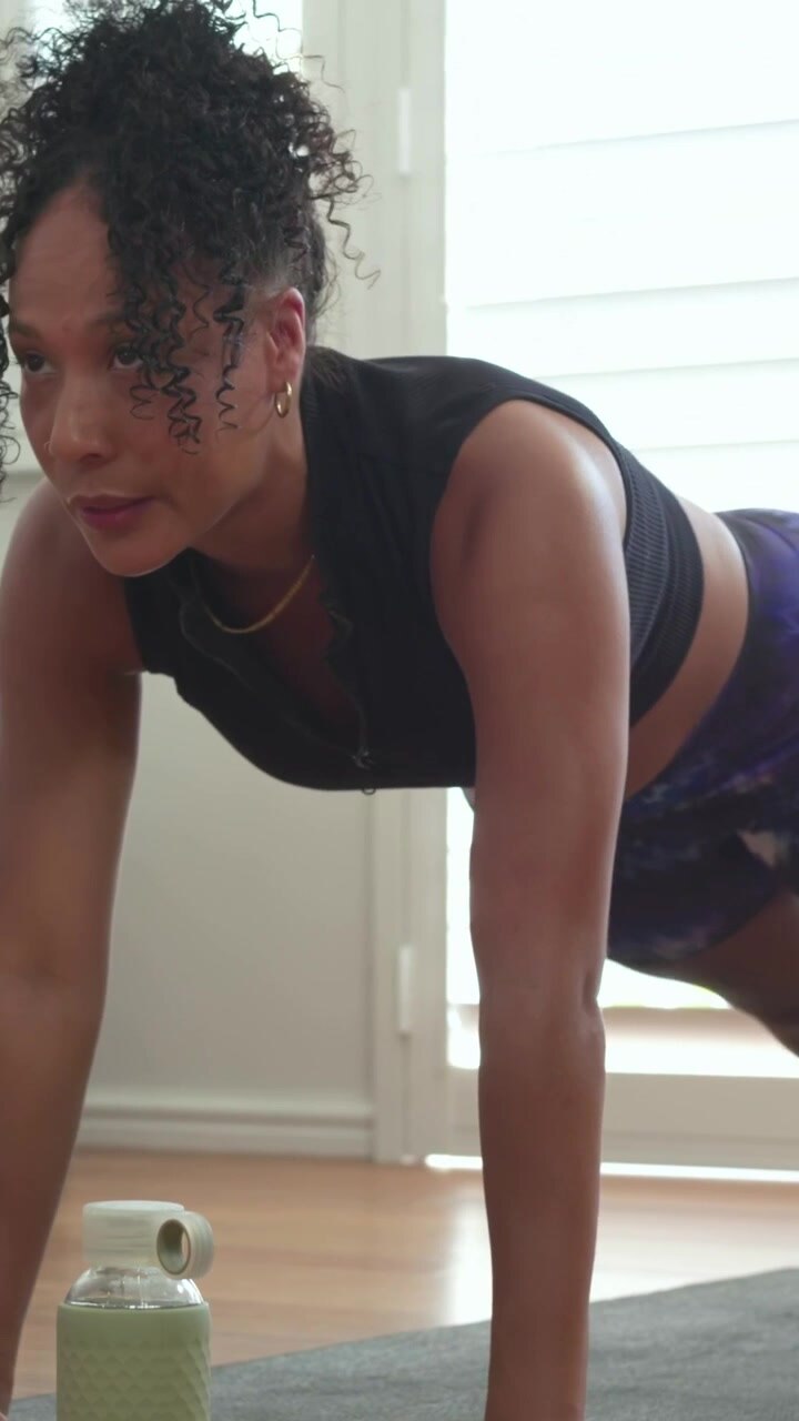 Vertical video: Starting plank, woman black top driving knees on home mat for core, water bottle