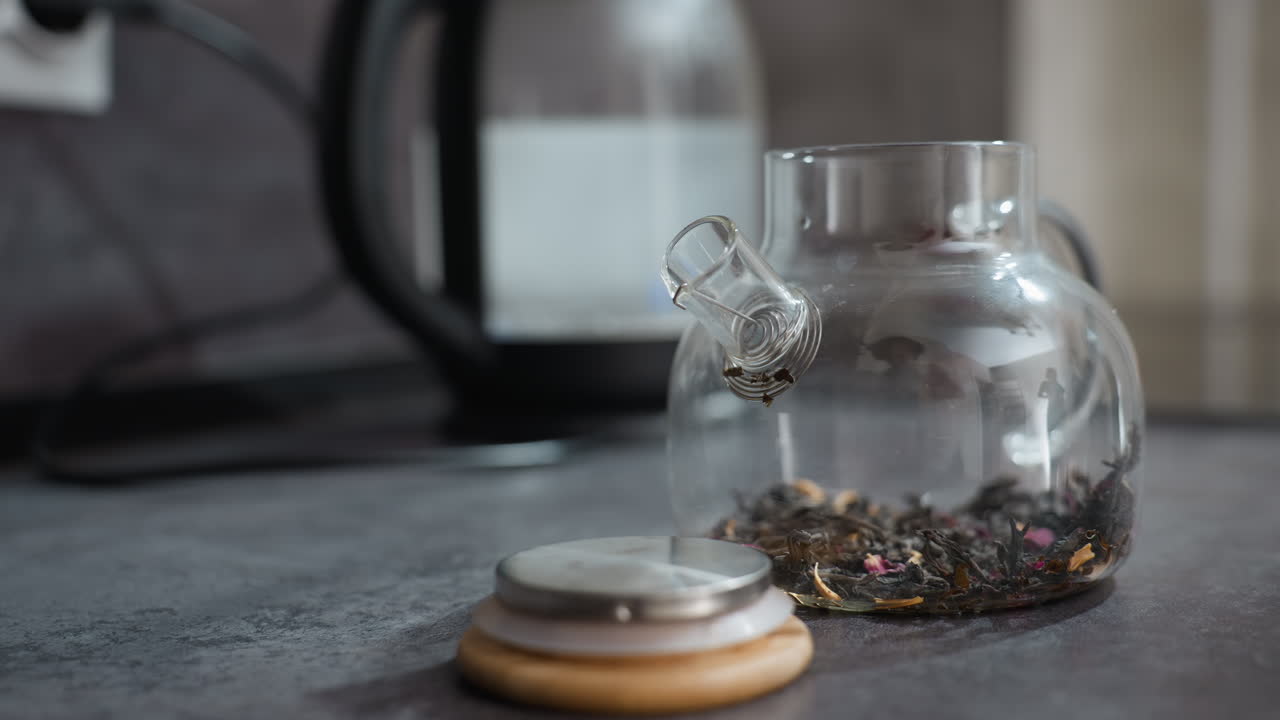 Glass Jar Of Dried Tea On Countertop With Kettle In Background, Warm Soft Light, Loose Leaves And Dried Berries Arranged Beside Lid, Calm Morning Ritual For Wellness And Cozy Kitchen Atmosphere