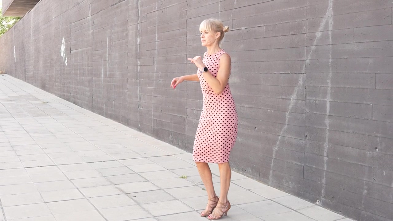 Uma mulher usando um vestido rosa está mandando um beijo e dançando ao ar livre em um cenário urbano.