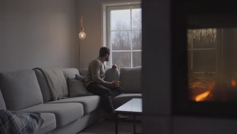 man relaxing in his living room, with a lit fireplace, looking out the window sipping a cup of tea