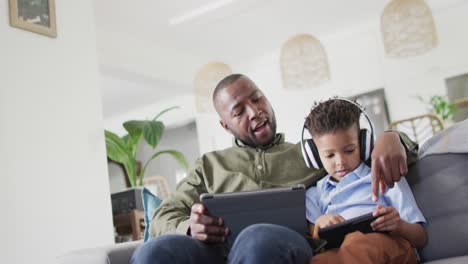 Happy-african-american-man-and-his-son-sitting-on-sofa-and-using-tablet