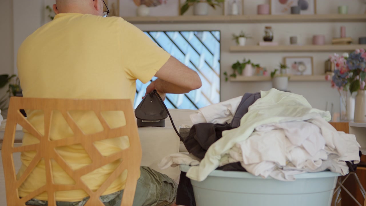 Back view of a man in a yellow shirt ironing clothes on an ironing board at home, with a large laundry basket full of unsorted clothes nearby, showcasing daily domestic chores and household tasks