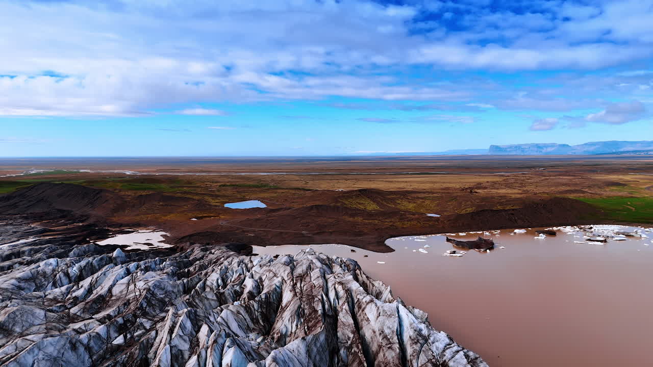Lake with brown water is between the ancient glacier and barren landscape. Amazing azure sky with some clouds is above the scenery. Iceland from top view.