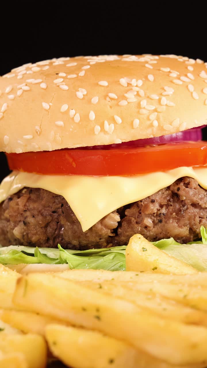 Un succulento cheeseburger di manzo, patatine fritte dorate e anelli di cipolla croccanti ruotano su una superficie riflettente sotto una brillante illuminazione da studio su uno sfondo nero.