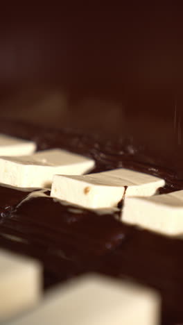 milky bars being covered by thick chocolate. white sweets moving under the flow of chocolate. production of candies close up. vertical video