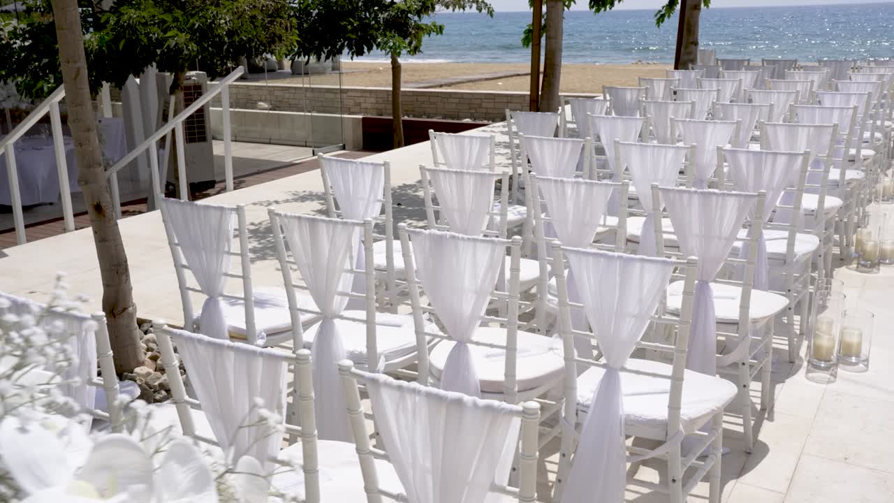 File di sedie bianche con drappeggi fluenti sono disposte per un matrimonio sulla spiaggia. La splendida vista sull'oceano aggiunge un tocco sereno all'ambiente romantico mentre iniziano i preparativi