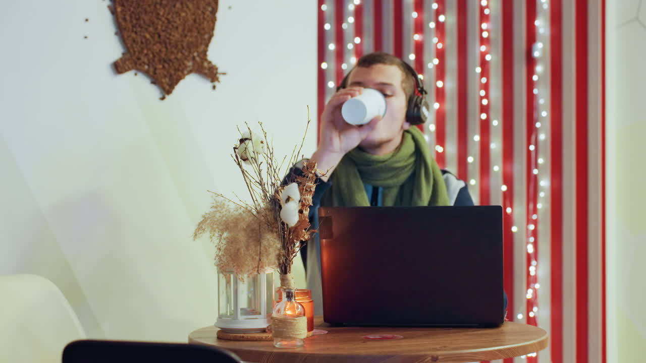 Giovane uomo caucasico che beve caffè al laptop, cuffie e sciarpa verde, vaso con fiori secchi, candela e lucine, digitazione concentrata e sorso occasionale, spazio di lavoro remoto accogliente per creativi.