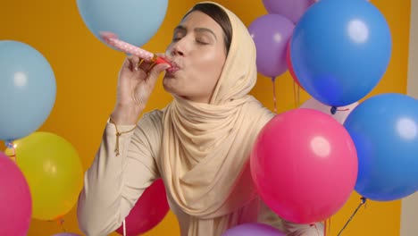 studio portrait of woman wearing hijab celebrating birthday with balloons and party blower