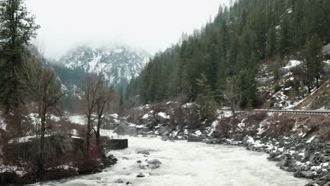 Cascade-Mountain-Pass-to-Leavenworth,-Washington---A-beautiful-Bavarian-styled-alpine-village-covered-in-snow---Rushing-River-Alpine-scene---Car-driving-by-in-distance