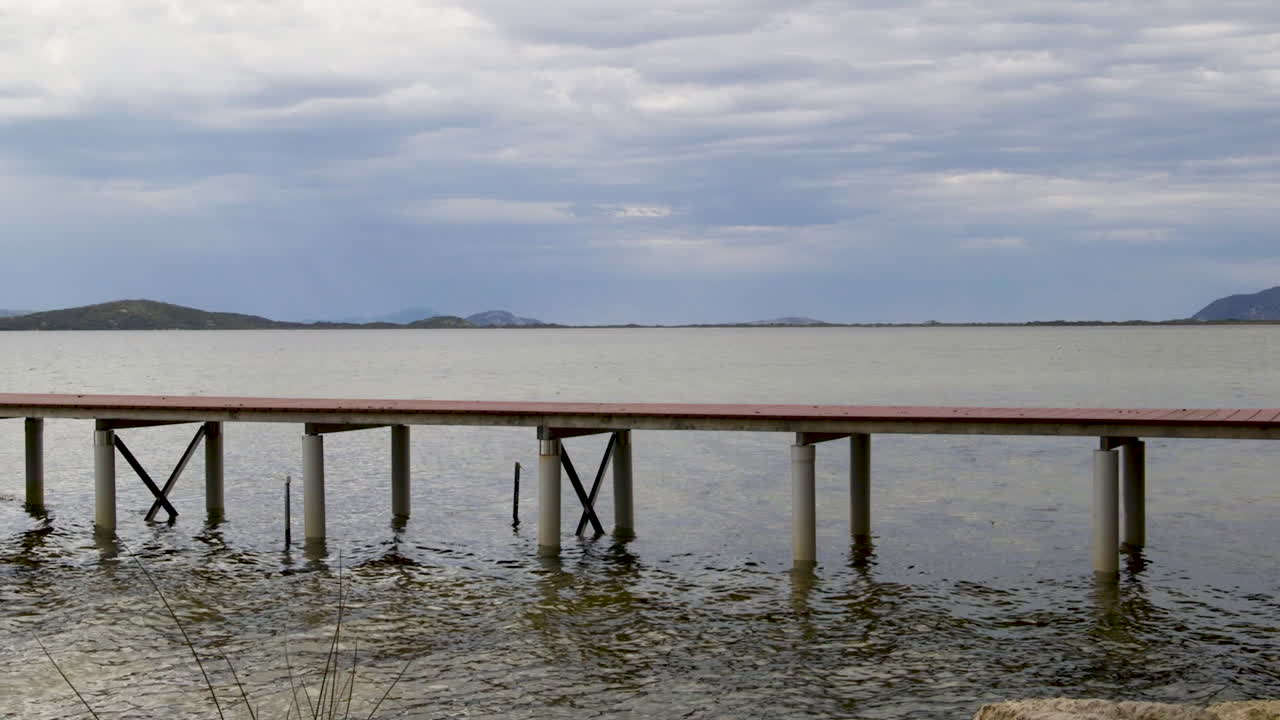 Un pontile si estende su acque calme, offrendo una vista sulle dolci colline sullo sfondo. L'immagine cattura l'ambiente naturale con un'illuminazione soffusa. Ideale per temi naturalistici o progetti sul lungomare.