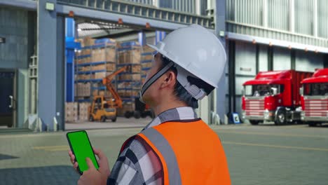 close up back view of asian male engineer with safety helmet working on a green screen smartphone and looking around while standing , outside of logistics distributions warehouse