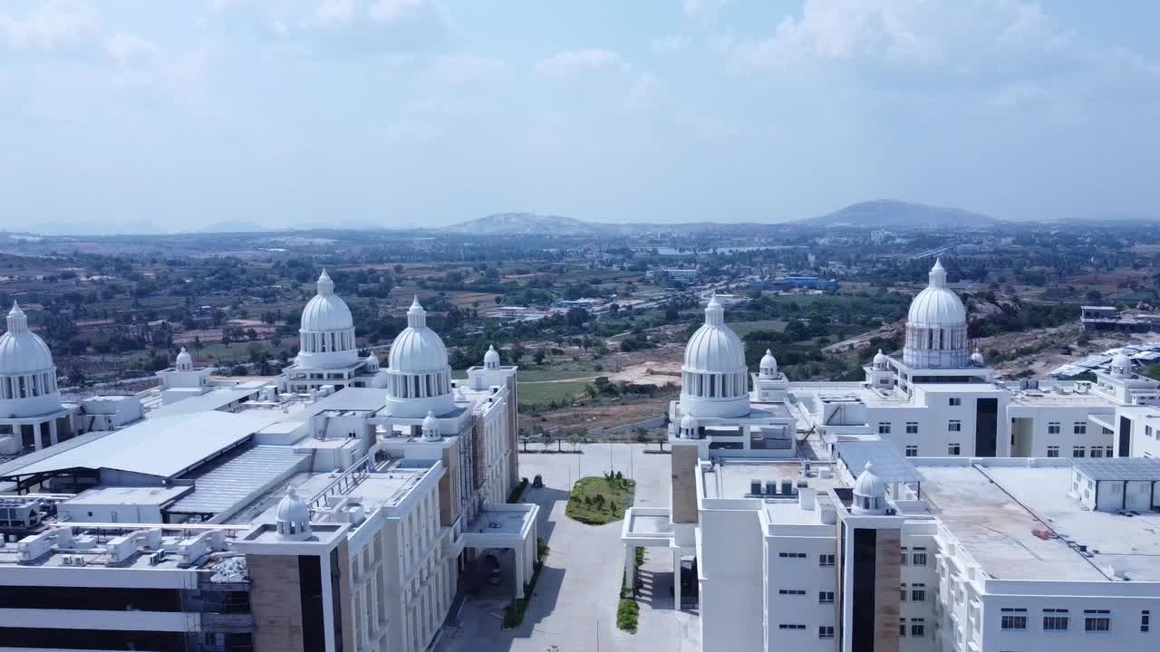 Wide aerial tracking shot showcasing the enormous, modern university or corporate campus with multiple white domes against a rural-urban fringe and distant hills