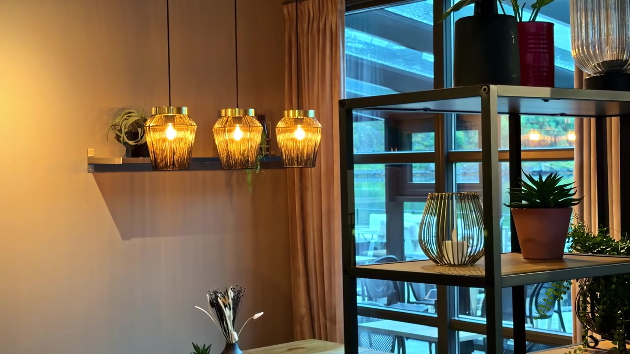 Lampu gantung hangat dan tanaman dalam ruangan menghiasi interior kafe modern yang nyaman di dekat jendela