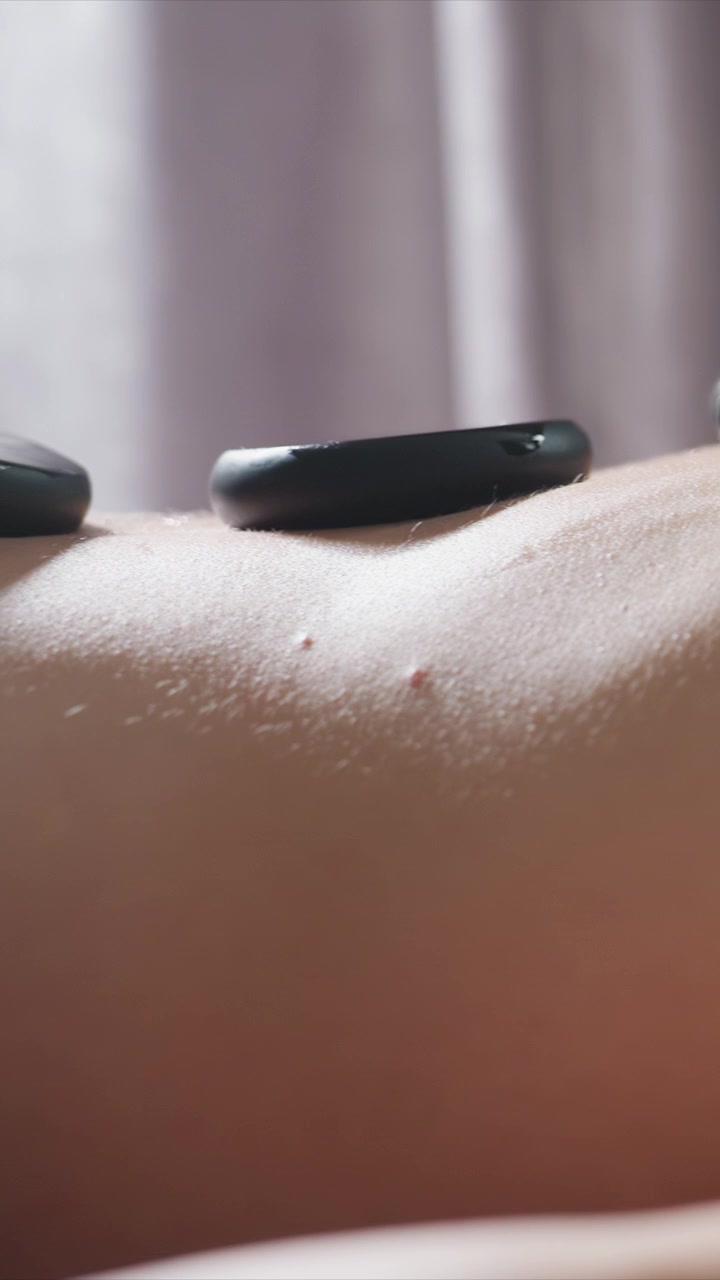 Nærbillede af en menneskeryg under en hot stone-massagebehandling. Glat hudtekstur er synlig, mens sorte sten placeres og fjernes, efterfulgt af en blid aftørring